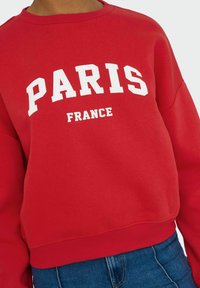 Rode sweatshirt met "PARIJS FRANKRIJK" gedrukt in vette witte letters. Het heeft een ronde hals, lange mouwen en ribgebreide boorden en onderkant.