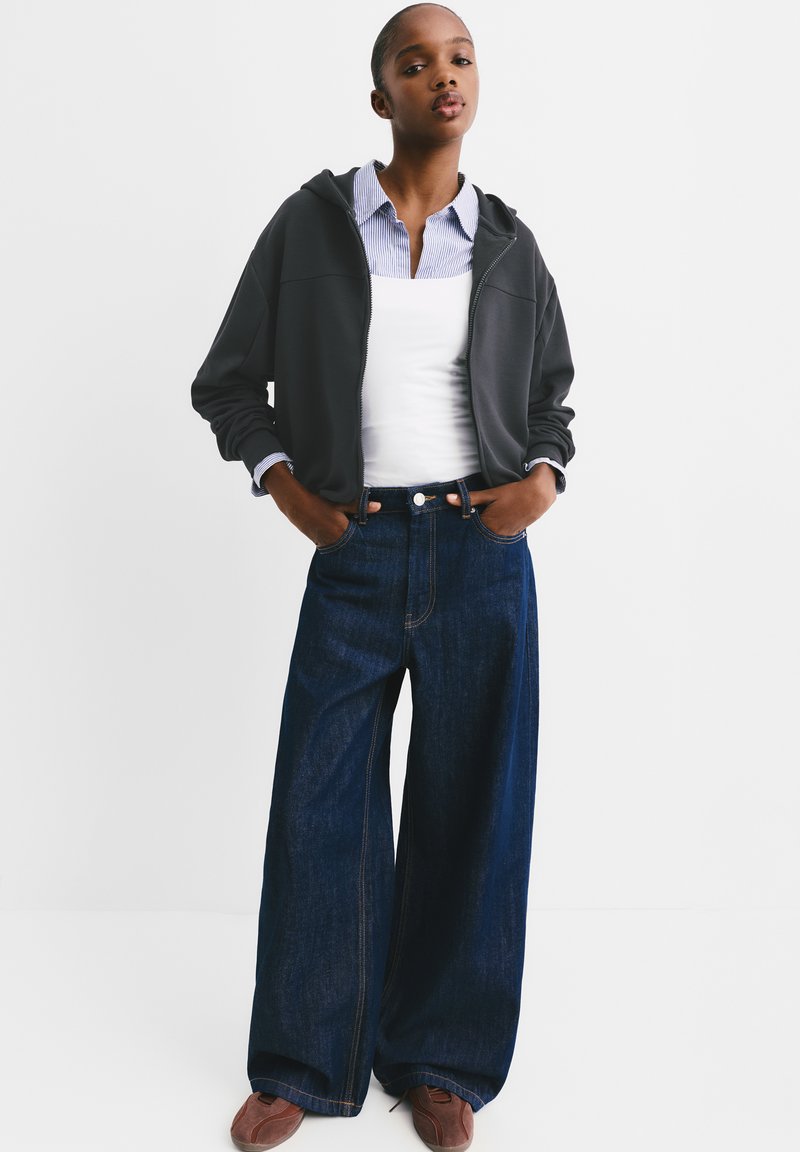 Dark zip hoodie worn open over a white fitted top with dark blue wide-leg jeans