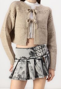 Béžový pletený krátký cardigan s dvěma mašlemi vp ředu, nošený přes bílý top. V kombinaci s skládacími sukní v černobílém tie-dye vzoru, která má několik spon.