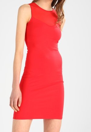 Femme portant une robe rouge ajustée sans manches avec un panneau transparent au niveau du décolleté, debout devant un fond clair uni.