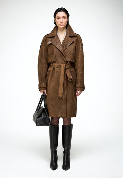 Žena stojaca, oblečená v hnedom semišovom trenčkotkovi so zaväzovaním v páse, čiernych čižmách po kolená a držiaca čiernu kabelku po boku.