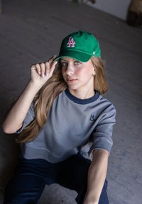 Groene honkbalpet met roze "LA" logo, gedragen door een persoon met lang haar. Draagt een gestreepte grijze en blauwe top. De scène is binnen.