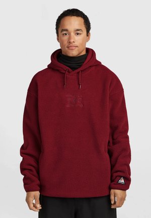 Man draagt een bordeauxrode fleece hoodie met trekkoorden en een zwarte coltrui eronder, staand tegen een effen lichte achtergrond.