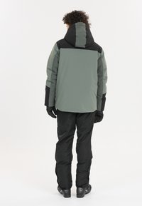 Hættejakke til vinter i grøn og sort, med et strømlinet design, struktureret stof og slidstærke handsker, parret med sorte bukser og støvler.