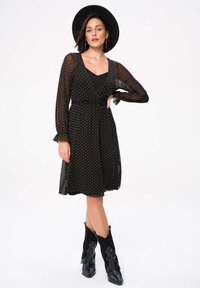 Zwarte jurk met stippen, lange doorschijnende mouwen, een geaccentueerde taille en een uitlopende rok. Gemaakt met een zwarte hoed en zwarte cowboylaarzen.