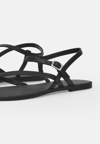 Black flat sandals featuring thin, crisscross straps made of textured leather, adjustable ankle buckle, and a smooth flat sole.