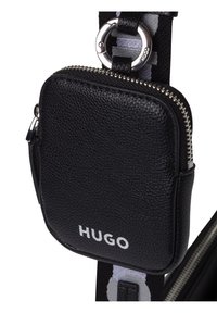 Funda de cuero negro texturizado con cremallera y herrajes plateados, cuenta con un logotipo en blanco en la parte frontal. Adjunta a una correa de tela a rayas.