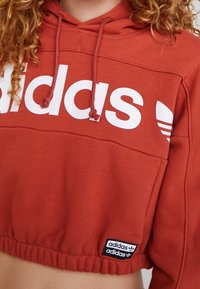 Kortärmad huvtröja i terrakotta med vitt "adidas"-logotyp. Har dragsko i huvan och elastisk nederkant, med en svart logotag på sidan.
