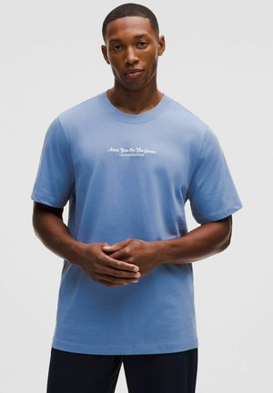 Homme portant un T-shirt bleu clair avec un texte blanc "Meet You On The Green, Lululemon Golf Club", mains jointes devant.