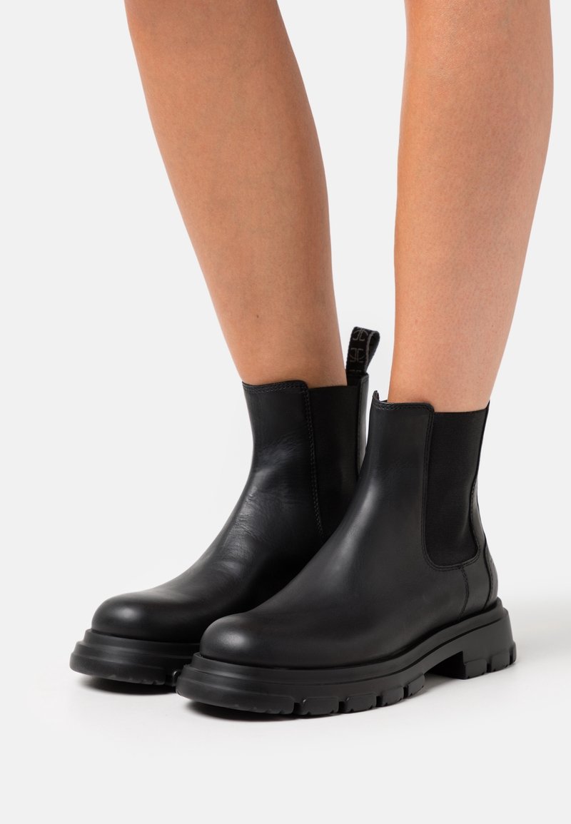 Bottes chevilles en cuir noir avec semelles épaisses portées sur des jambes nues, sur un fond blanc uni.