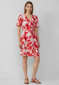 Robe à motifs rouges et roses avec des manches courtes, décolleté en V, taille cintrée et jupe fluide. Portée avec des sandales marron. Texture de tissu lisse.