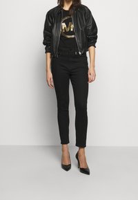 Svart läderbomberjacka med samlade ärmslut, tillsammans med en svart t-shirt med en guldglittrig logotyp och svarta skinny jeans och klackar.