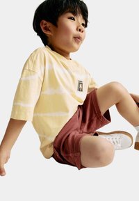 Camiseta amarilla con efecto tie-dye y mangas cortas, combinada con pantalones cortos de color óxido. Zapatillas blancas con suelas de goma. Diseño minimalista y ajuste casual.