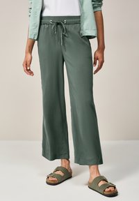 Pantalons légers, amples, de couleur vert foncé, avec une taille élastique et un cordon de serrage. Associés à des sandales à glissière de couleur vert olive.