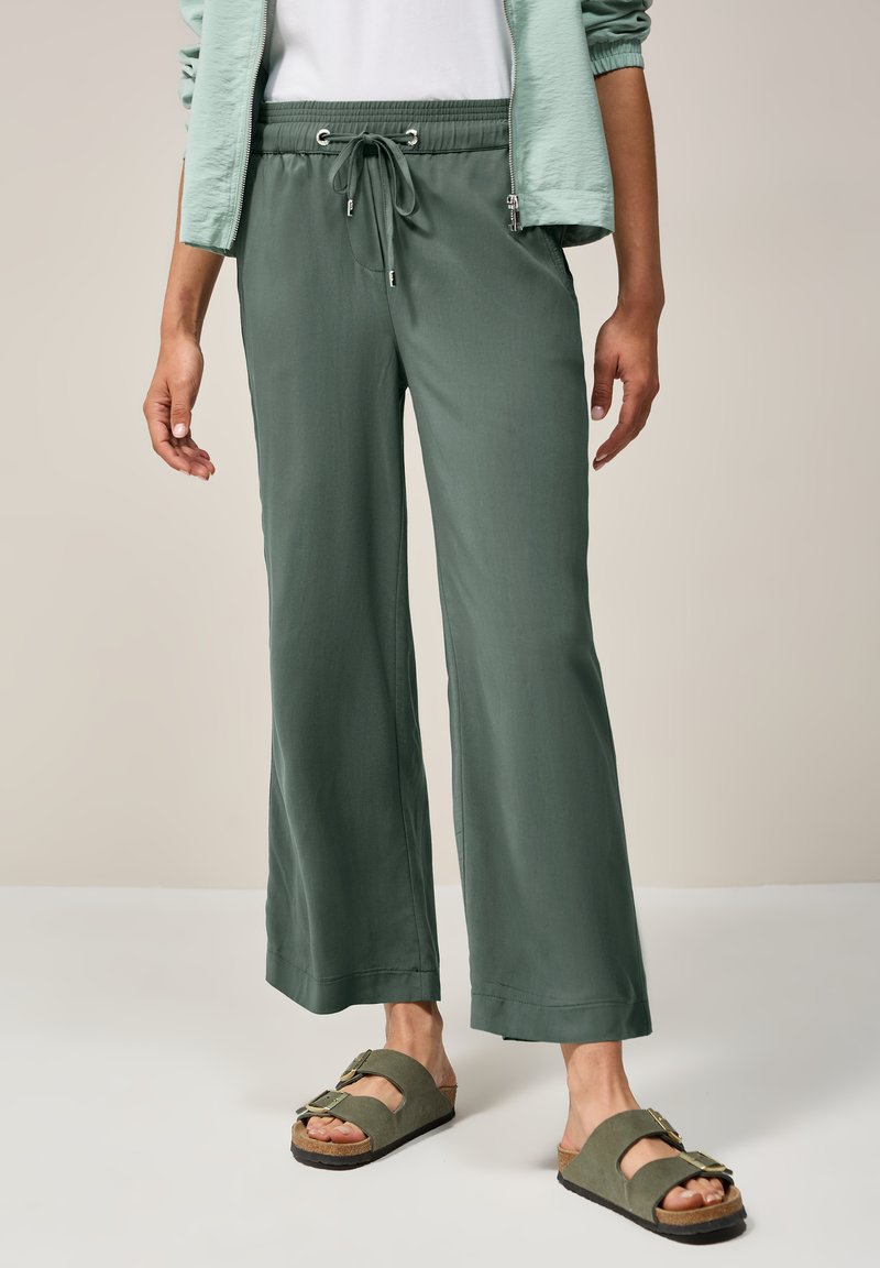 Pantalons légers, amples, de couleur vert foncé, avec une taille élastique et un cordon de serrage. Associés à des sandales à glissière de couleur vert olive.