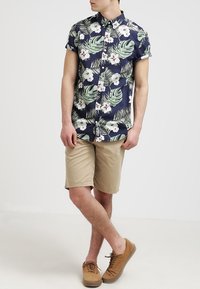 Krátký rukáv námořnicky modré košile s bílými květinovými a zelenými listovými vzory, doplněný béžovými šortkami. Outfit je doplněn světle hnědými botami.