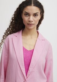 Pink linen blazer with a notched collar, worn over a bright pink knit top. Curly hair and neutral background. Soft texture and tailored design.