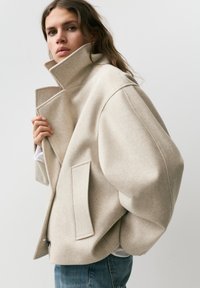 Kobieta ubrana w oversize'owy beżowy wełniany płaszcz z wysokim kołnierzem i niebieskie dżinsy, patrząca w stronę aparatu na jednolitym tle.