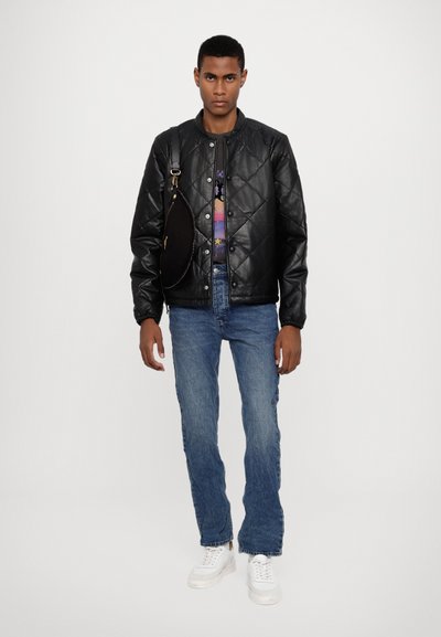 Veste en cuir matelassée noire, jeans bleus et baskets blanches. La veste est dotée de fermetures à pression ; la tenue comprend une chemise à motifs en dessous.