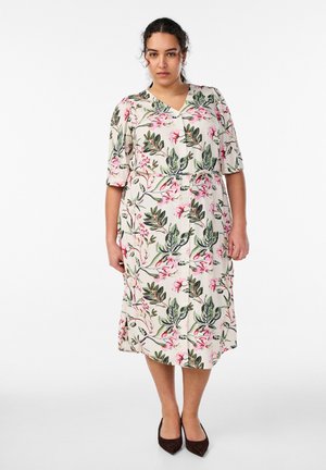 Naine, kes kannab põlveni ulatuvat nööpidega kreemikat kleiti rohelise ja roosaka lillemustriga ning mustade teravate varbaga kingadega, seisab valge tausta ees.