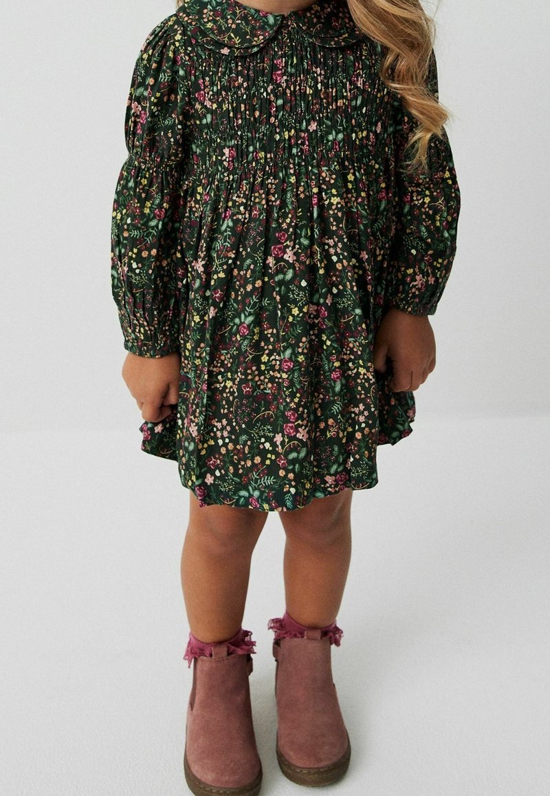 Enfant portant une robe fleurie vert foncé à manches longues et des bottines roses, debout sur un sol clair.