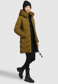 Giacca puffer verde oliva con chiusura a zip, motivo a chevron e cappuccio. Abbinata a leggings neri e stivaletti. Design semplice e elementi funzionali.