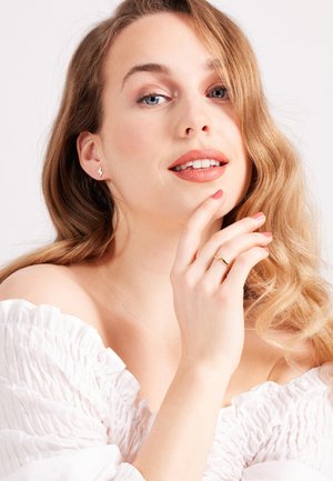 Ein Model mit welligem, blonden Haar trägt goldene Blitze Ohrringe und einen dünnen goldenen Ring, gekleidet in einem strukturierten, weißen Off-Shoulder-Oberteil.