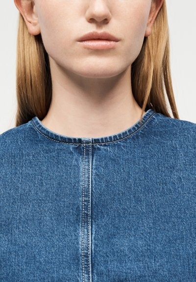 Κοντινό πλάνο ενός ατόμου με ίσια ξανθά μαλλιά που φοράει μπλε τοπ από τζιν με εμφανείς ραφές κατά μήκος της λαιμόκοψης και της μπροστινής ραφής.