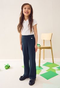 Pantalones de pierna ancha de color verde oscuro con cintura alta y un pequeño emblema floral. Combinados con una camisa polo blanca de manga corta y zapatos negros.