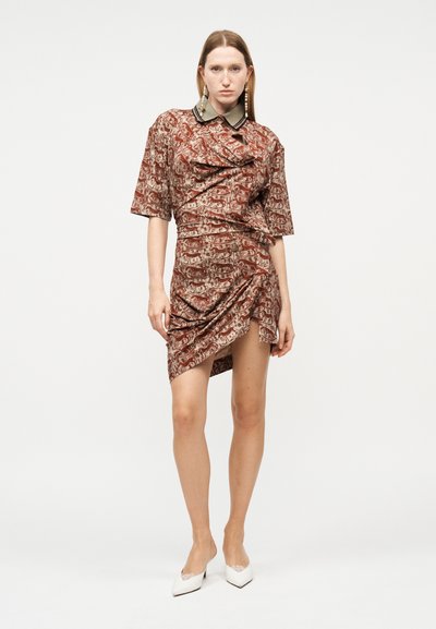 Kvinde iført en brun kort kjole med mønster og grøn krave, med puffede og rynkede detaljer, kombineret med hvide spidse hæle.