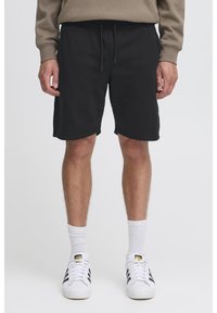 Shorts en coton noir avec une taille élastique et un cordon de serrage ajustable. Texture lisse, longueur au genou, portés avec des chaussettes blanches et des baskets.