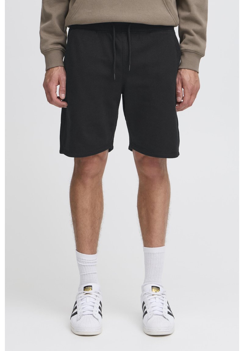 Shorts en coton noir avec une taille élastique et un cordon de serrage ajustable. Texture lisse, longueur au genou, portés avec des chaussettes blanches et des baskets.