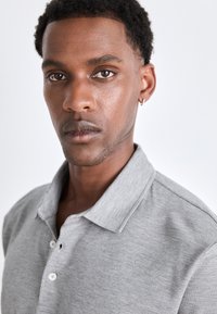 Pilkas polo marškinėliai, pagaminti iš minkštos medžiagos, turi sulankstomą apykaklę ir tris sagas. Artimiausias modelio veido planas su trumpais plaukais.