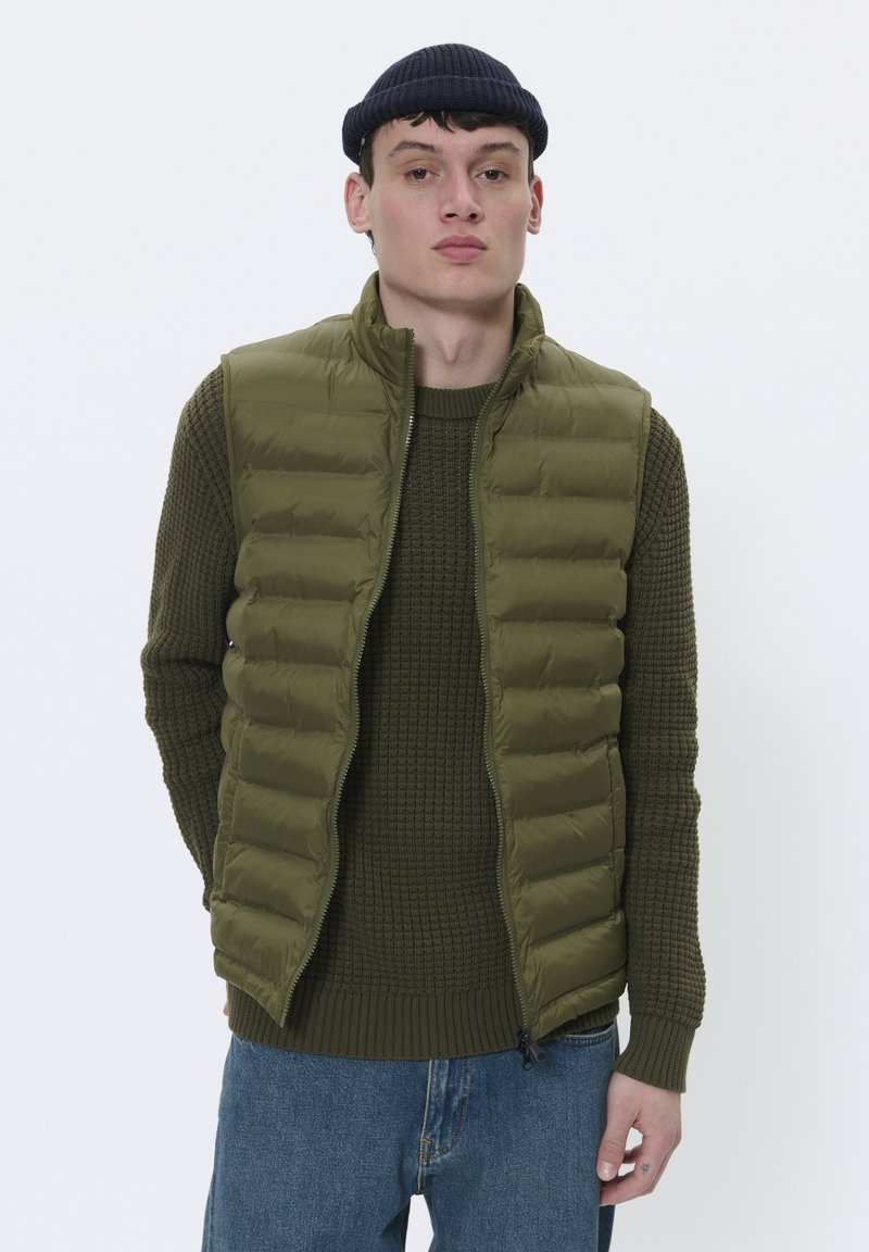 Prsluk od maslinaste zelene boje s patentnim zatvaračem i visokim ovratnikom, nosi se preko teksturirane zelene veste. Model stoji protiv jednostavne bijele pozadine.