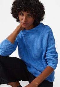 Maglione blu lavorato a maglia con texture a coste, scollatura ampia e maniche lunghe leggermente arrotolate. Abbinato a pantaloni neri.