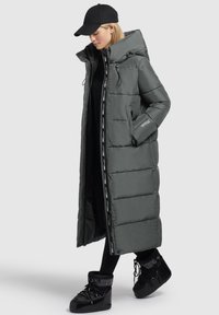 Lange grijze pufferjas met een hoge kraag en ritszakken, voorzien van een zwarte binnenvoering en logo-accenten aan de zijkanten. Gecombineerd met zwarte schoenen.