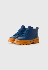 Botas de tornozelo em couro azul com solas de borracha laranja grossas e atacadores, posicionadas lado a lado sobre um fundo branco liso.