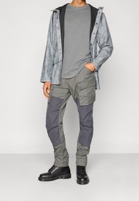 Szara, wodoodporna kurtka o teksturowanym wykończeniu, szary t-shirt pod spodem oraz dwukolorowe spodnie cargo; całość dopełniają czarne buty.