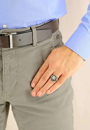 Anello nero con un design di teschio poggia su una mano, con pantaloni grigi e una cintura marrone. Camicia azzurro chiaro visibile sopra la cintura.