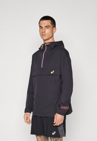 Leichte schwarze Jacke mit einem halben Reißverschluss, Kapuze und Fronttasche. Mit bunten Zugbändern und Logo-Akzent. Kombiniert mit schwarzen Shorts.