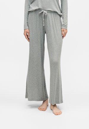 Femme portant un pantalon ample gris avec un cordon à la taille et un haut gris à manches longues assorti, pieds nus sur fond blanc.