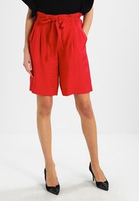 Rote hochgeschnittene Shorts mit weitem Bein, Stoffgürtel und seitlichen Taschen, die eine glatte Textur und einen lockeren Schnitt haben, kombiniert mit schwarzen spitzen Absätzen.