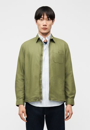 Mladý muž s rovnými čiernymi vlasmi, oblečený v olivovozelenej bunde cez svetlú pruhovanú košeľu na gombíky, stojaci pred bielym pozadím.