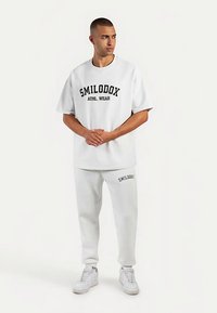 Vitt set i bomull för träning med kortärmad topp med texten "SMILODOX ATHL. WEAR" och matchande mjukisbyxor med logotypdetalj.