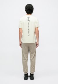 Krēmkrāsas īsām piedurknēm T-krekls ar vertikālu melnu logotipa apdruku uz muguras. Kombinēts ar dūmakainiem biksēm un melniem apaviem.