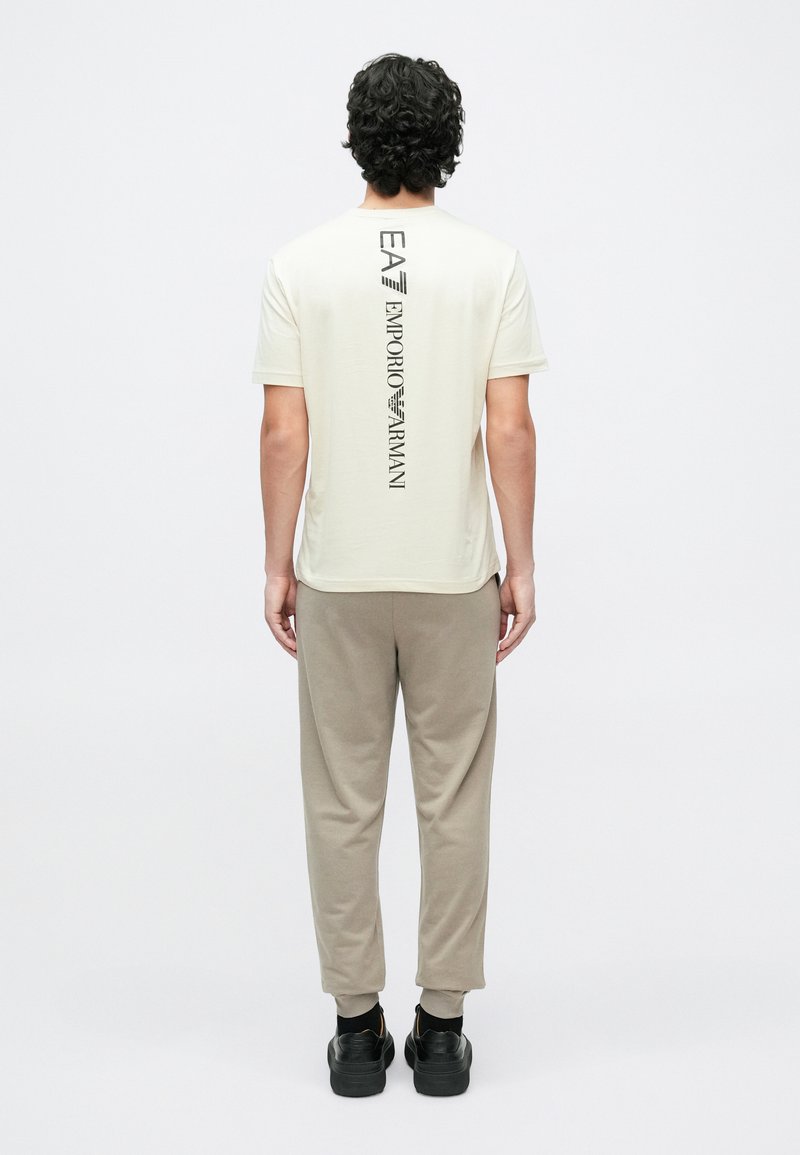 Krēmkrāsas īsām piedurknēm T-krekls ar vertikālu melnu logotipa apdruku uz muguras. Kombinēts ar dūmakainiem biksēm un melniem apaviem.