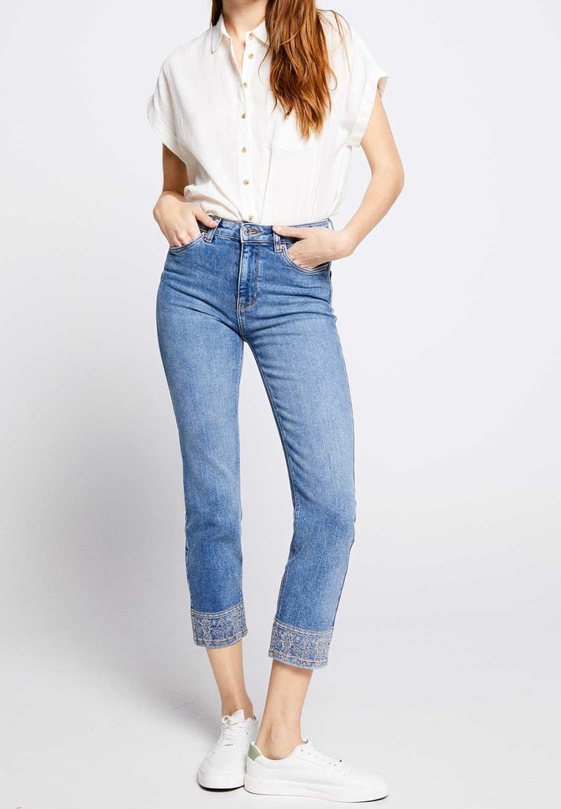 Kvinna klädd i en vit kortärmad skjorta med knappar, instoppad i högmidjade jeans avkortade i blått med broderade ärmslut och vita sneakers.