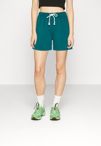 Teal puuvillased lühikesed püksid elastse vöökoha ja valge kootud nööriga, kombineeritud roheliste spordijalatsite ja valgete lühi-sokkidega.