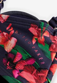 Sejas maska no auduma ar ziedu rakstu, kurā spilgti rozā un sarkanas ziedi uz tumši zila fona, centrā redzams zīmols.