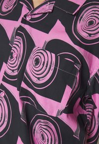 Lyhythihainen nappipaita mustasta kankaasta, jossa on vaaleanpunaisia ympyrä- ja geometrisia kuvioita, rintatasku ja vaaleanpunaiset napit.